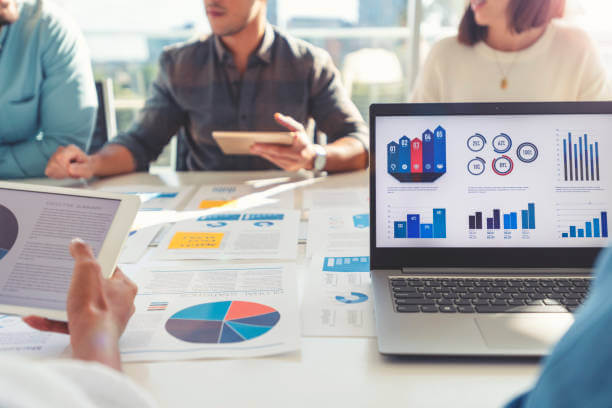 Group of people meeting with technology and paperwork. Close up of digital tablet and laptop computer with market analysis data on the screen. The documents have financial or marketing figures, graphs and charts on them. Multi ethnic group including Caucasian, latin and African American