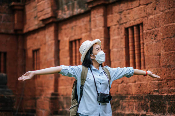 Young asian backpacker female wearing hat and protection mask while traveling in historic site, she use camera take a photo with happy