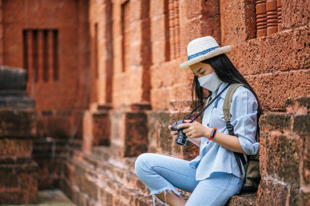 Young asian backpacker female wearing hat and protection mask while traveling in historic site, she checking photo in camera with happy