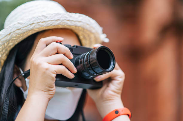 Young asian backpacker female wearing hat and protection mask while traveling in historic site, she use camera take a photo with happy