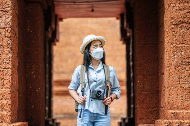 Young asian backpacker female wearing hat and protection mask while traveling in historic site, she use camera take a photo with happy