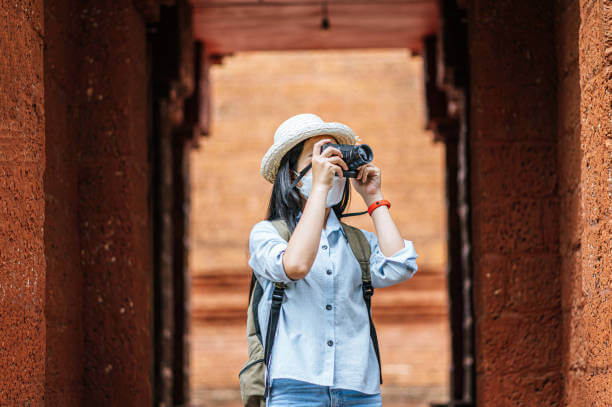 Young asian backpacker female wearing hat and protection mask while traveling in historic site, she use camera take a photo with happy