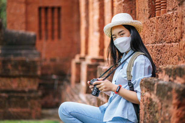 Young asian backpacker female wearing hat and protection mask while traveling in historic site, she use camera take a photo with happy