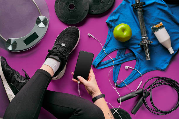 Young woman with mobile phone and accessories for sports, bottle of water, headphones, fitness bracelet, dumbbells on color background flat lay top view. Fitness concept