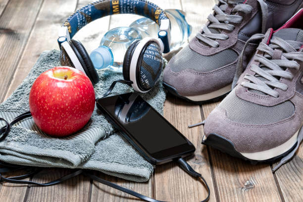 Close-up of a red apple, sport shoes, audio headphone, smartphone, towel and water bottle photographed on an antique wooden table.
