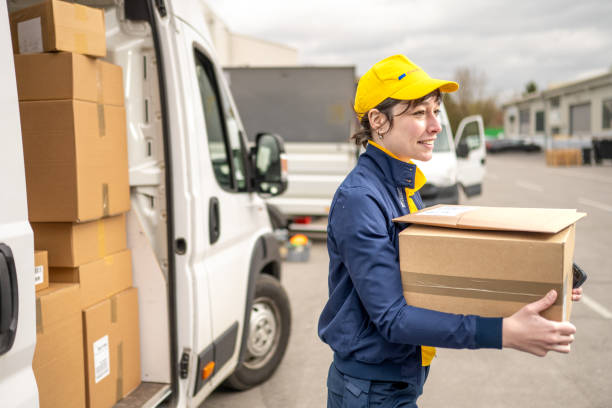 Young female courier in unifrom carrying carboard box, unloading delivery truck. Shipment service, postal worker holding order package outdoors, side view medium shot