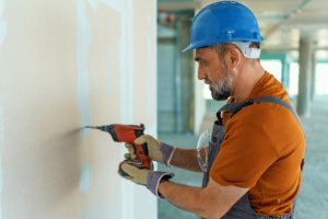 A mature Caucasian male construction worker is standing on a construction site and using an electric drill.