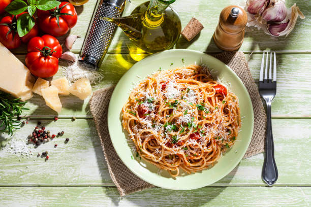 Top view of traditional Italian pasta plate and ingredients for cooking on green kitchen table. A cheese grater an a wooden pepper mill are visible in the composition The ingredients included are fresh organic tomatoes, basil leaves, olive oil, peppercorn, garlic, Parmesan cheese and raw spaghetti. DSRL studio photo taken with Canon EOS 5D Mk II and Canon EF 100mm f/2.8L Macro IS USM