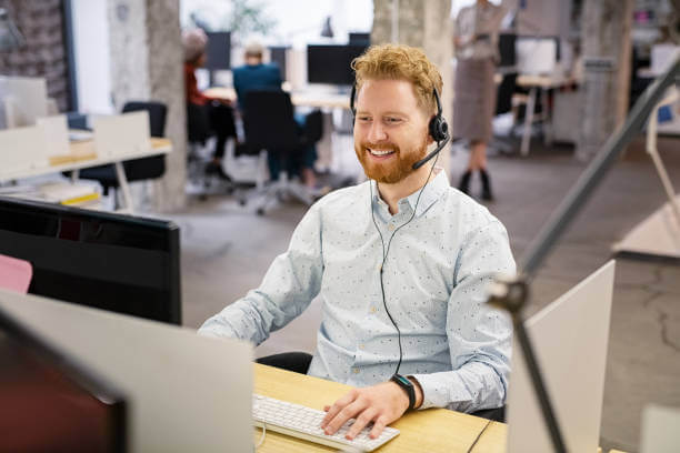 Portrait of consultant agent man in call center smiling. Happy customer support agent working with headset while sitting at his workstation. Smiling telephone operator using computer in modern open space office.