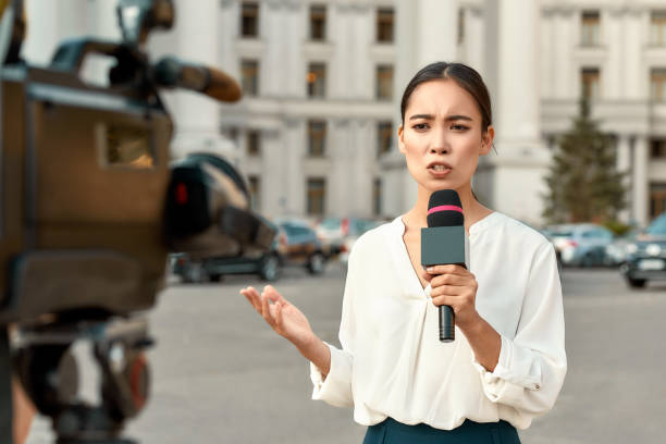 Cropped portrait of professional female reporter at work. Young woman standing on the street with a microphone in hand and smiling at camera. Horizontal shot. Selective focus on woman