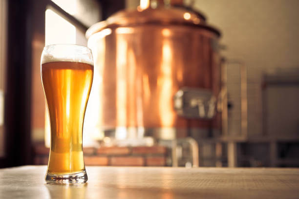 Glass of lager beer on the table in the microbrewery. Copper vat in the background. No people.