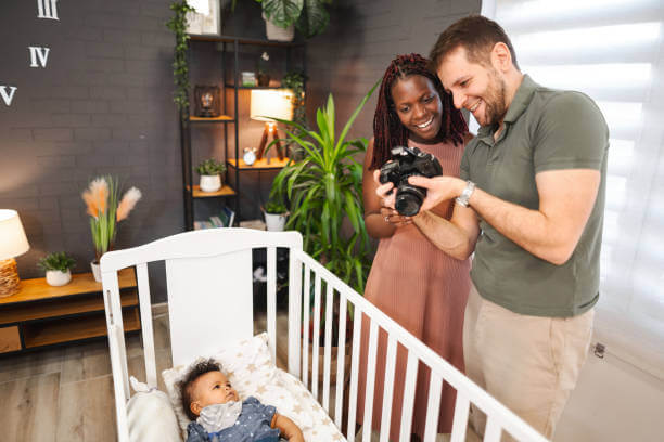 Multiracial married couple taking pictures with professional camera of their newborn baby while lying in nursery bed