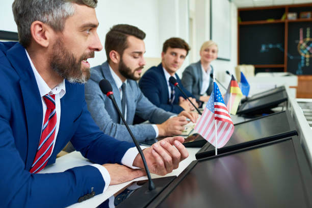 Portrait of several business people sitting in row participating in political debate during press conference focus on bearded man answering media questions speaking to microphone