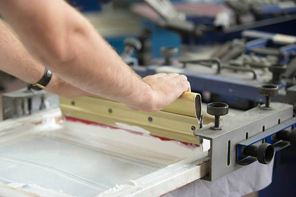 Close up of male hands exploring traditional squeegee, creating an image on white fabric clothing by pressing ink through a screen with areas blocked off by a stencil