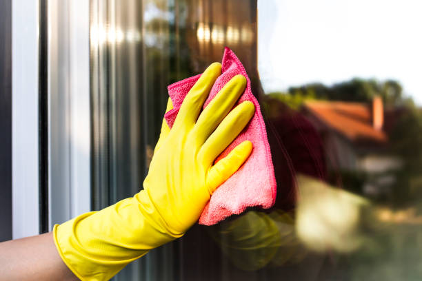 A hand in yellow protective glove cleaning the glass window pane with a pink rag. Home routine.