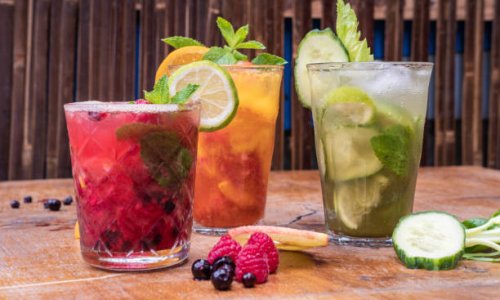 Front view of 3 fresh fruit juices in glasses on a wooden table, there are cucumber and peach slices on the table, with raspberries and blueberries, glasses garnished with mint leaves, lime and orange slices.