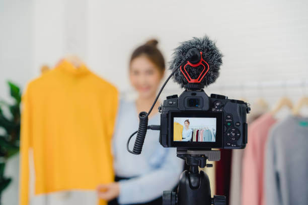 Asian fashion female blogger online influencer holding shopping bags and lots of clothes on clothes rack for recording new fashion video broadcast live video to social network by internet at home.