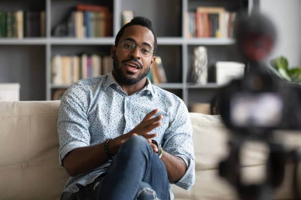 Millennial african hipster man blogger recording vlog on digital camera sit on sofa in living room, confident young guy vlogger influencer shooting social media video blog on camcorder talk at home